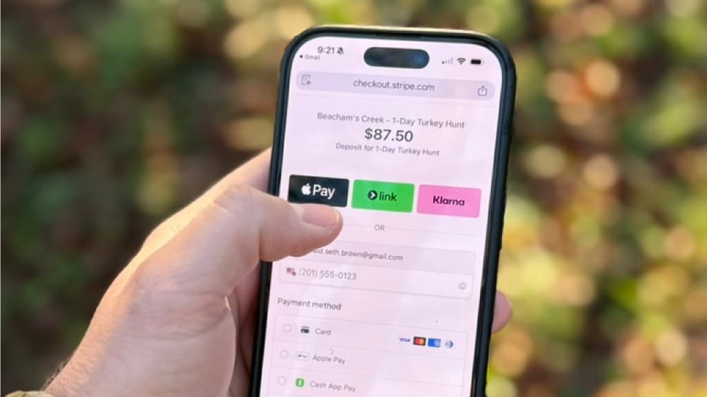Hand holding a smartphone outdoors showing a hosted checkout page for a hunting trip deposit